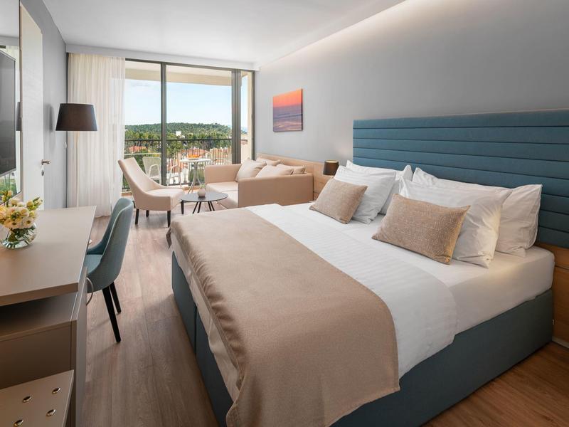 Modernes Hotelzimmer mit Doppelbett, Schreibtisch, Sessel und Balkon mit Aussicht auf Grünfläche.