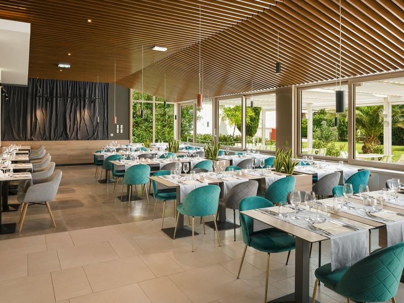 Modernes Restaurant mit türkisfarbenen Stühlen, großen Fenstern und Holzbalkendecke.