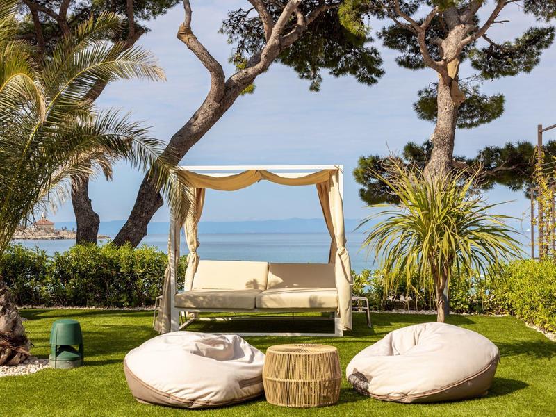 Garten mit grüner Wiese, Palmen, zwei Sitzsäcken, Holztisch und schattigem Tagesbett am Meer.