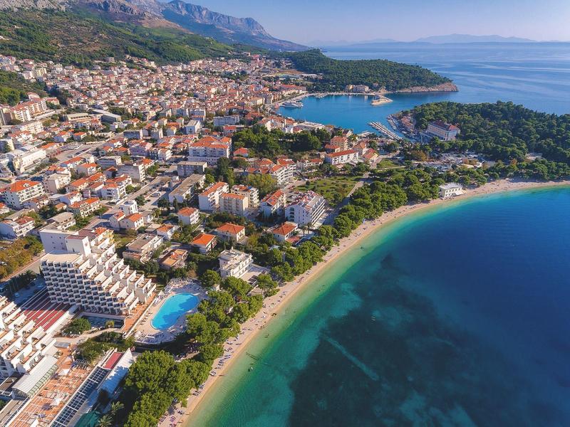 Städtische Küstenlandschaft mit Sandstrand, klarem Wasser, Gebäuden und bewaldeten Hügeln.