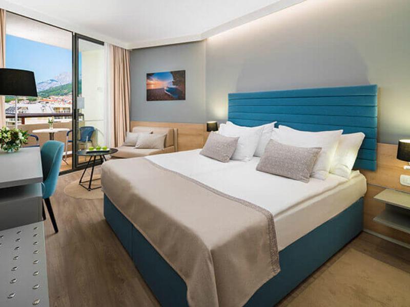 Modernes Hotelzimmer mit großem Bett, blauen Akzenten und einem Fenster mit Ausblick auf Berge.