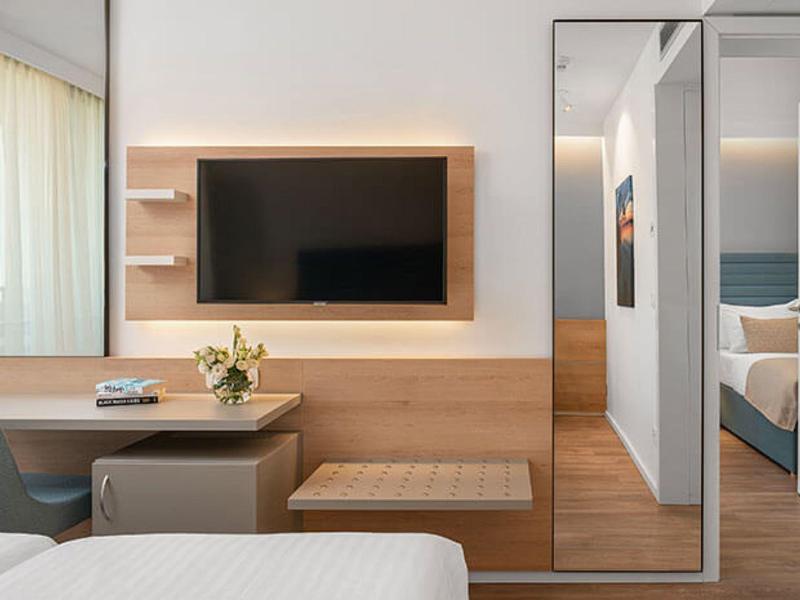 Moderner Hotelraum mit Wand-TV, Holzmöbeln, großem Spiegel und offenem Blick ins angrenzende Schlafzimmer.