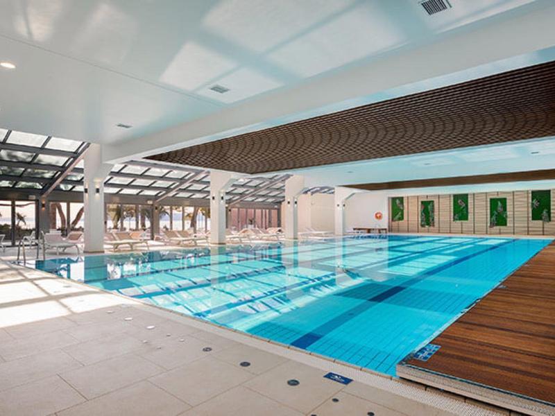 Hallenbad mit blauem Wasser, Holz- und Steinboden, weißen Wänden und großem Fenster für viel Licht.