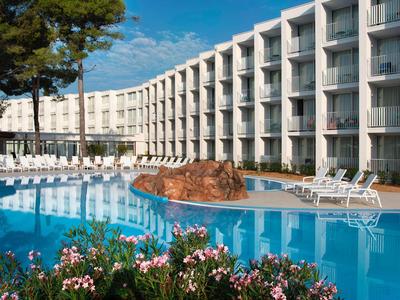 Großes Hotel mit geschwungenem Pool und Liegestühlen unter blauem Himmel.