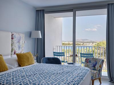 Helles Hotelzimmer mit Balkon, blau-gemustertem Bett und Blick auf eine Landschaft mit Hügeln.