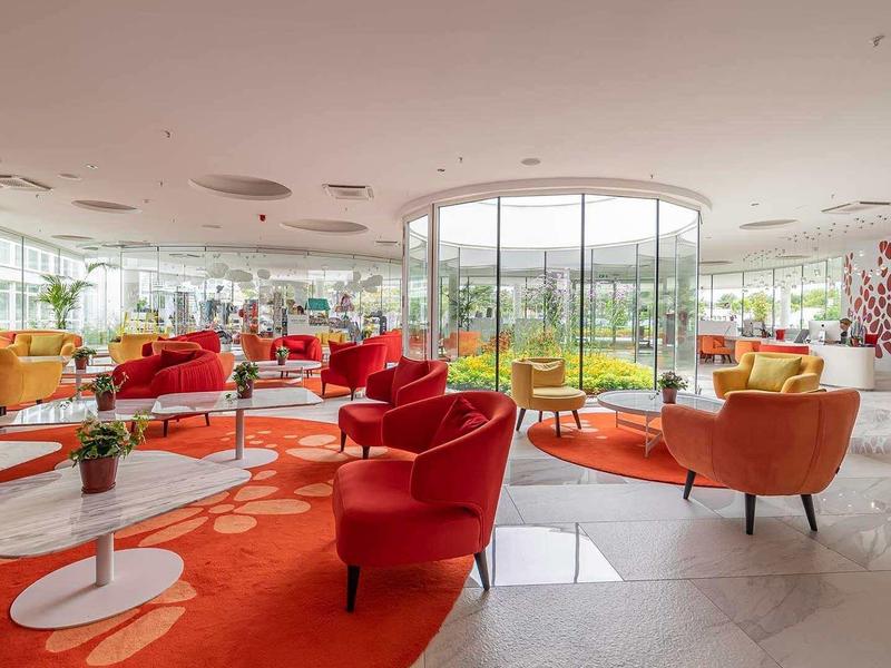 Heller Raum mit großen Fenstern, roten Sesseln, orangem Teppich und weißen Tischen.