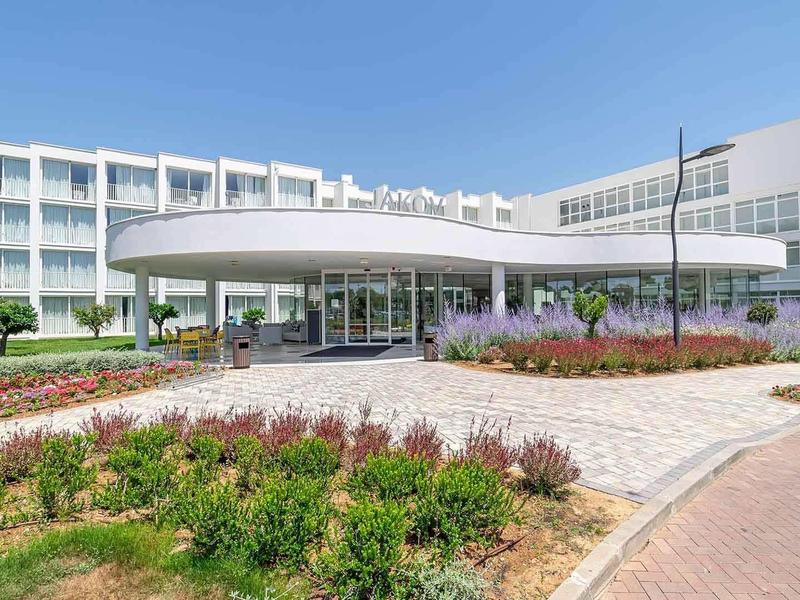 Modernes weißes Gebäude mit kreisförmigem Eingang, Blumenbeeten und blauem Himmel im Hintergrund.