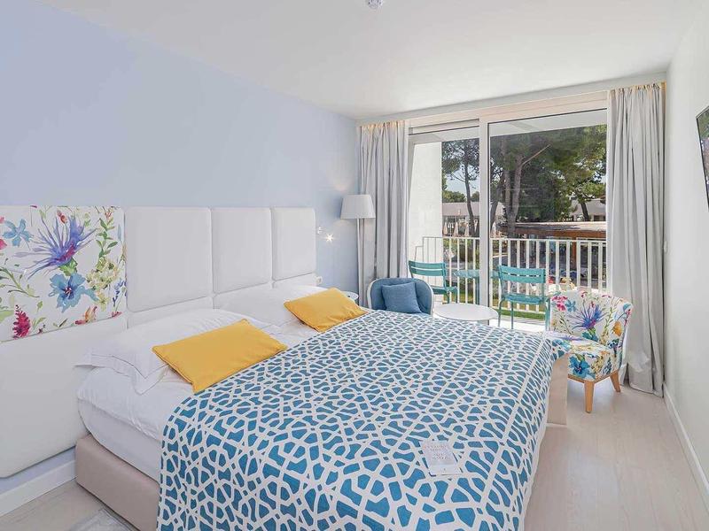 Helles Schlafzimmer mit großem Bett, gemusterten Decken, Balkon und modernem TV an der Wand.