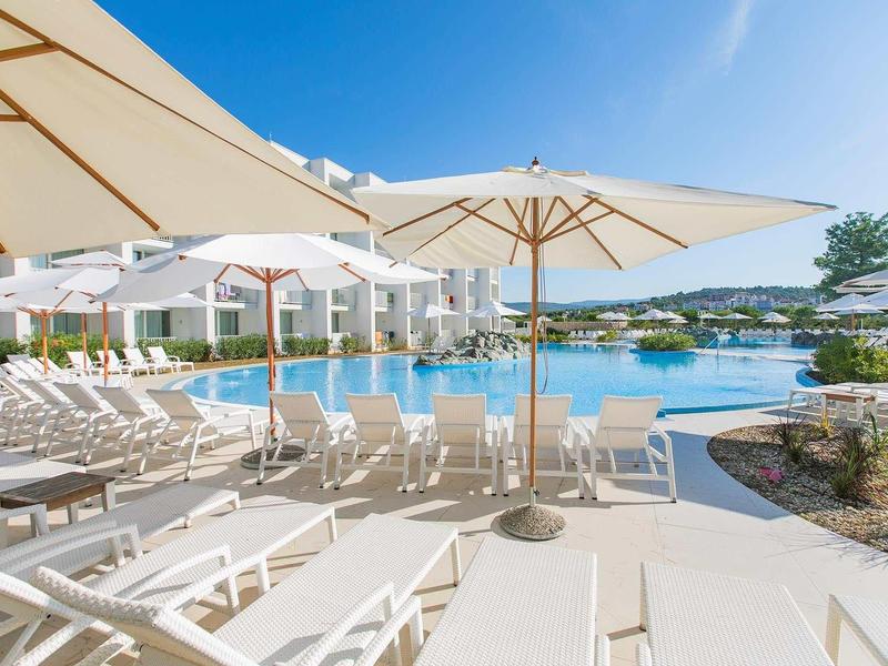 Poolbereich mit weißen Liegestühlen und großen Sonnenschirmen unter klarem blauem Himmel.