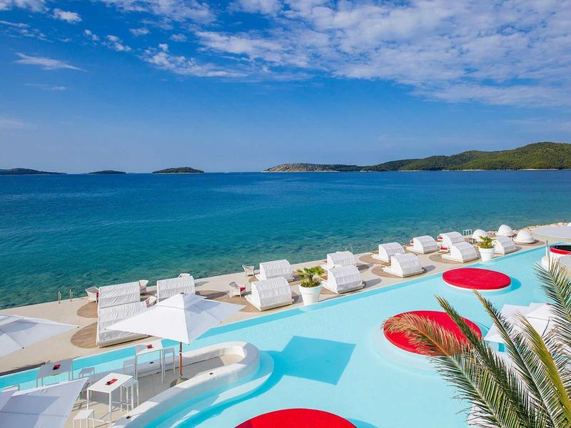 Strandhotel mit blauem Pool, weißen Liegen, Sonnenschirmen und Blick auf das ruhige Meer.