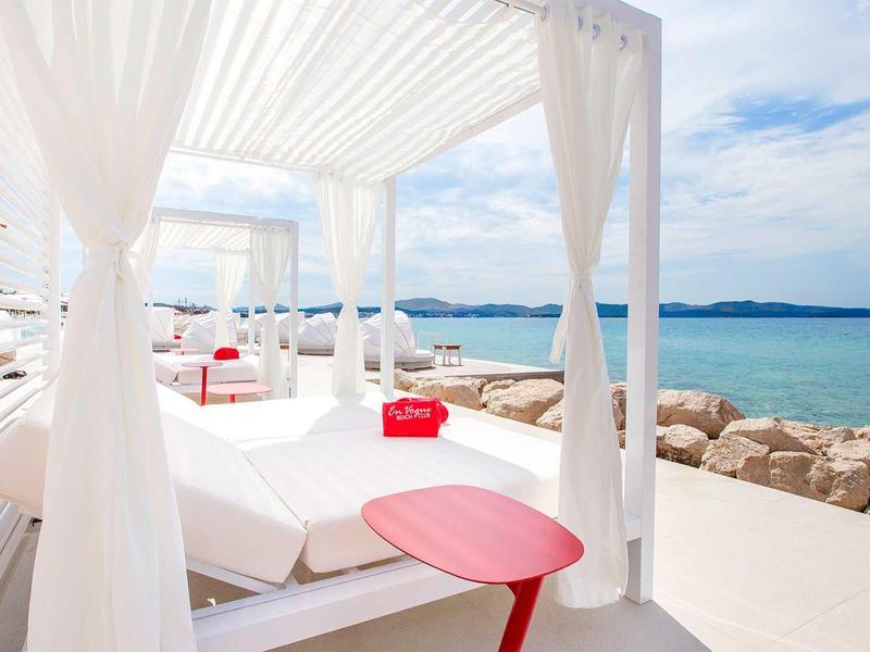 Weiße Strandliegen mit Vorhängen und rotem Tisch mit Blick auf das blaue Meer und den Himmel.