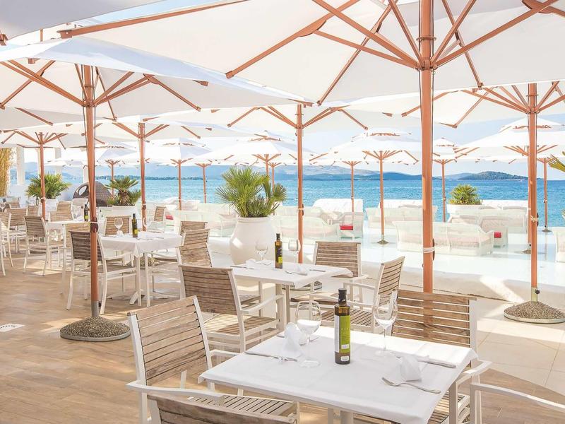 Terrassenrestaurant mit weißen Sonnenschirmen, Holztischen und Meerblick bei klarem Himmel.