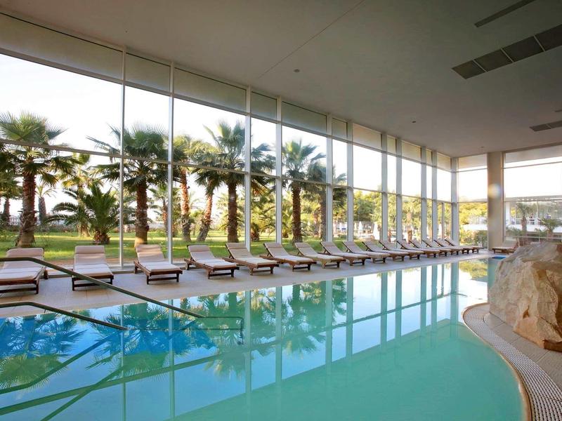 Großes Hallenbad mit Liegestühlen, Glaswänden und Blick auf Palmen im Außenbereich.