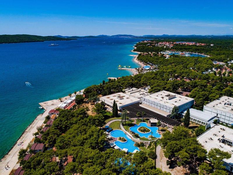 Küstenlandschaft mit blauem Meer, bewaldeten Hügeln und mehreren weißen Gebäuden mit Pools.