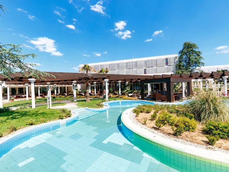 Ein Gebäude mit Sonnendach, umgeben von einem blauen Wasserlauf und grünen Pflanzen unter blauem Himmel.