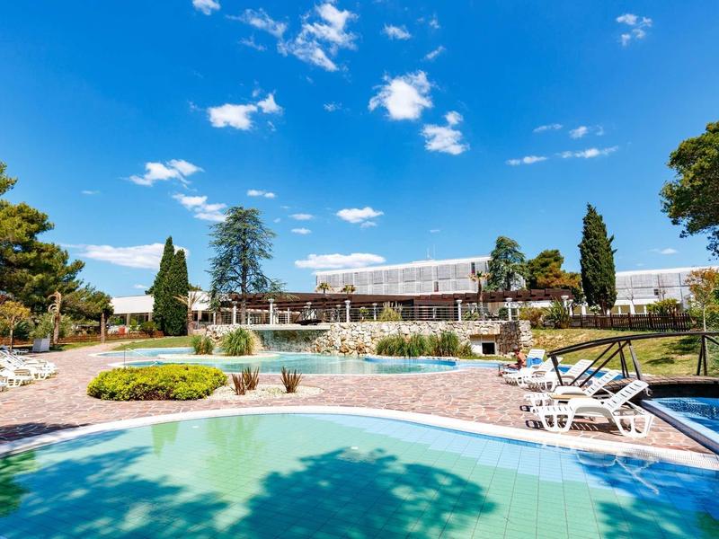 Großer Außenpool mit Liegestühlen, grünem Garten und bewölktem blauem Himmel im Hintergrund.