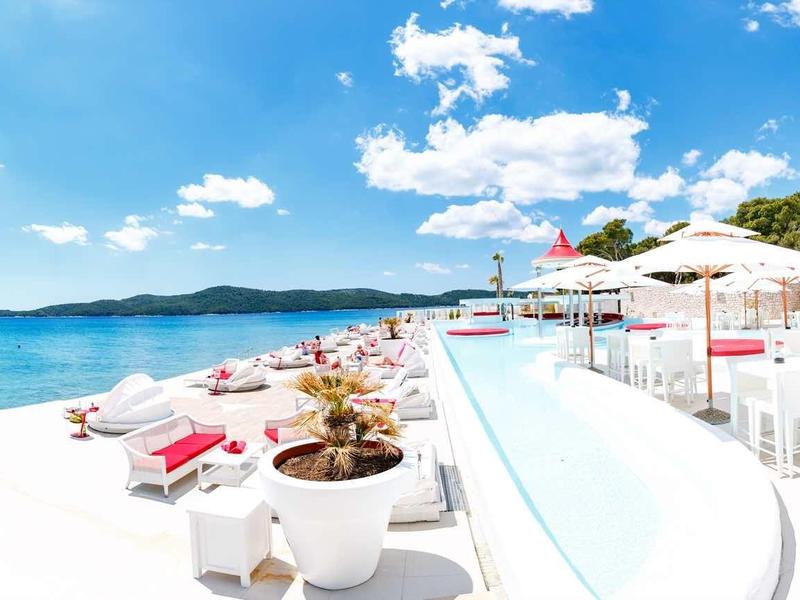 Heller, sonniger Strand mit weißem Sand, blauen Liegen, Pool und klarem Himmel mit wenigen Wolken.