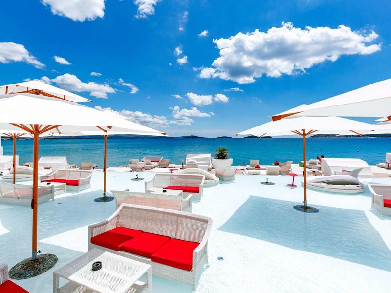 Strandbereich mit weißen Liegen, roten Polstern und Sonnenschirmen unter blauem Himmel und Wolken.