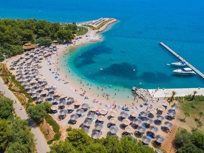 Strand mit vielen Sonnenschirmen, blauem Meer und Holzsteg, umgeben von grünen Bäumen.