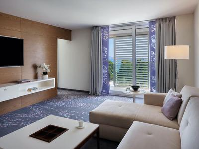 Heller Hotelzimmer-Wohnbereich mit Sofa, Teppich und Balkon mit Blick auf Grünflächen.