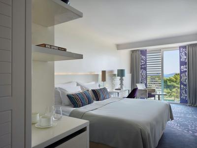 Helles Hotelzimmer mit großem Bett, Sitzbereich, geöffnetem Fenster und Blick ins Grüne.