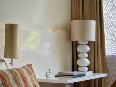 Moderne Hotelzimmer-Ecke mit beigem Bett, Streifenkissen und zwei Schreibtischlampen neben einem weißen Schreibtisch.