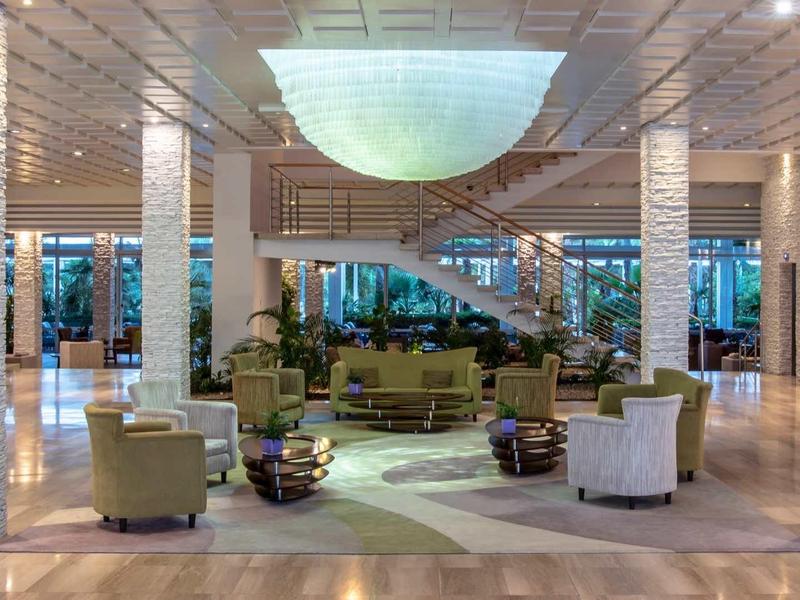 Großer heller Empfangsbereich mit weißen Säulen, Rundsofa, Sesseln und modernem Treppenaufgang.