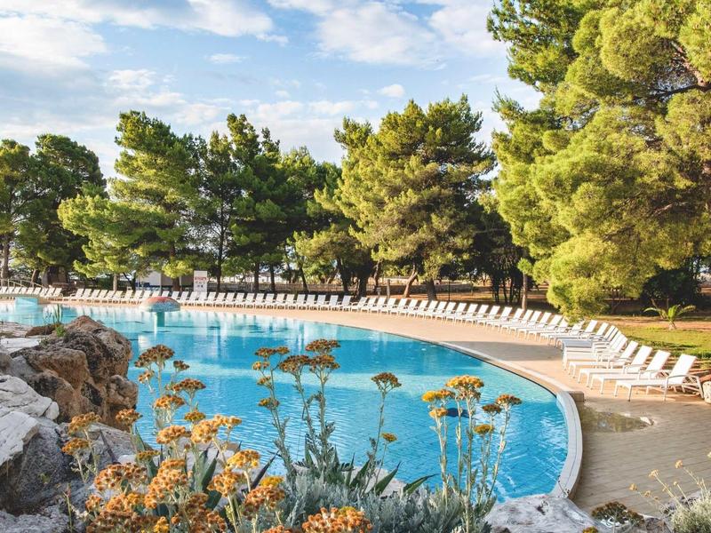 Rundes Freibad mit blauem Wasser, umgeben von Liegestühlen und Pinien unter blauem Himmel.