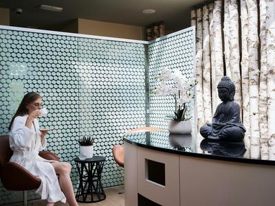 Frau im Bademantel sitzt in modernem Spa-Bereich mit Buddha-Statue und Pflanze.