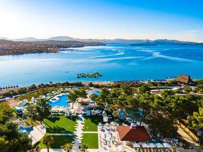 Vista de un resort con grandes piscinas junto a la orilla de un lago azul tranquilo bajo un cielo despejado.