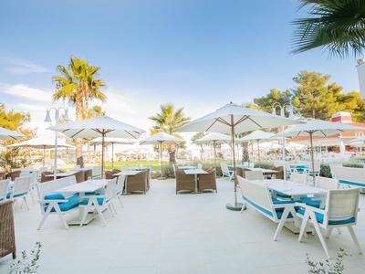 Restaurante en la playa con mesas blancas, sillas azules y sombrillas bajo palmeras.