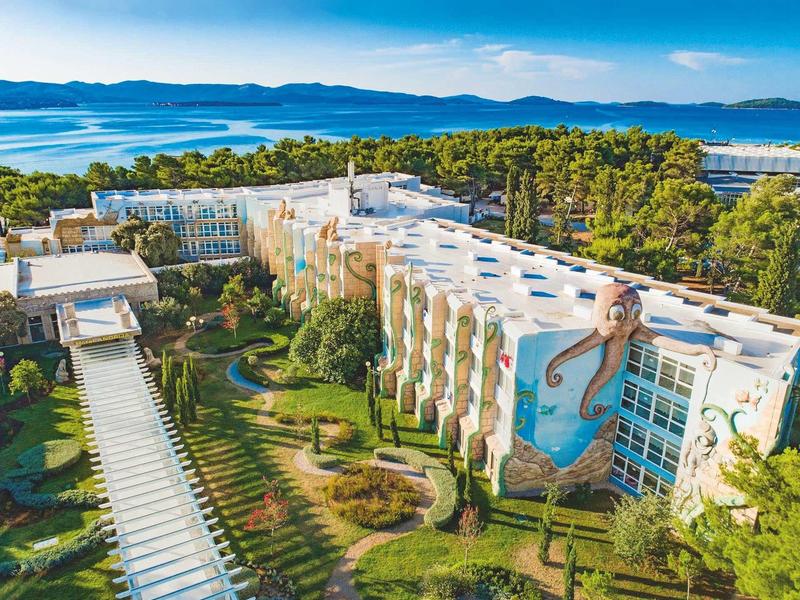 Groot hotel met muurschildering aan gevel, omgeven door bos en uitzicht op zee.