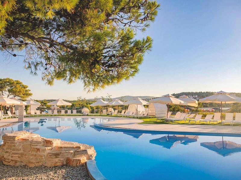 Groot zwembad met parasols en stoelen in een zonnig hotelgebied met bomen.