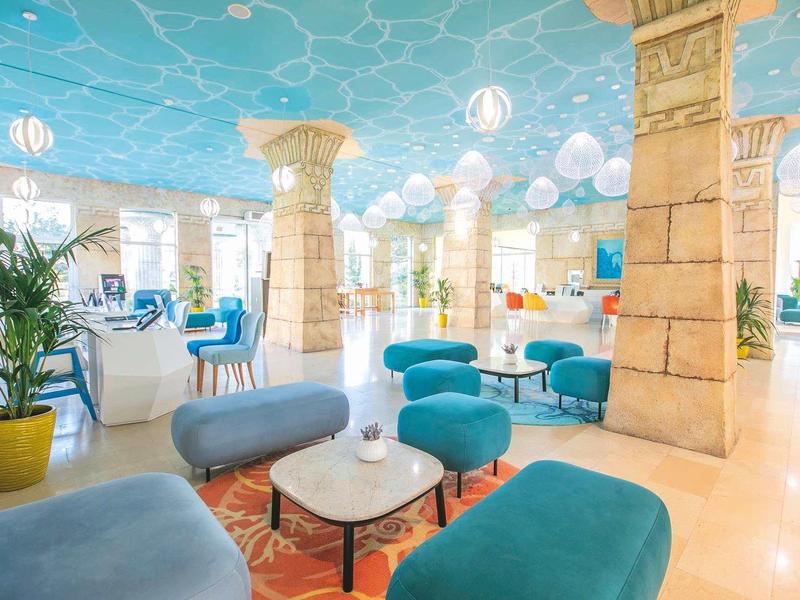 Lichte hotelinterieur met turquoise plafond, zuilen en blauwe en grijze zitgedeeltes.