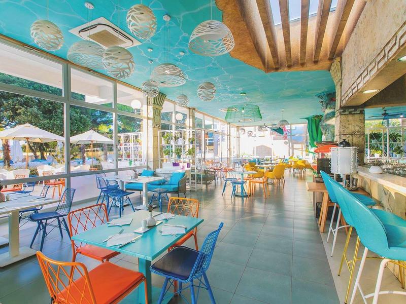 Lichte hotelbar met kleurrijke stoelen en grote ramen met uitzicht op parasols buiten.