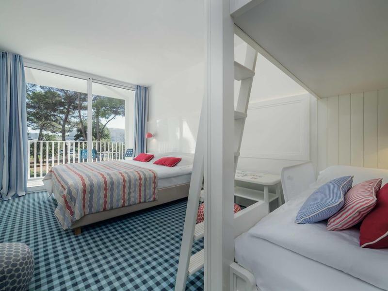 Lichte hotelkamer met tweepersoonsbed, stapelbed en balkon uitzicht met bomen.