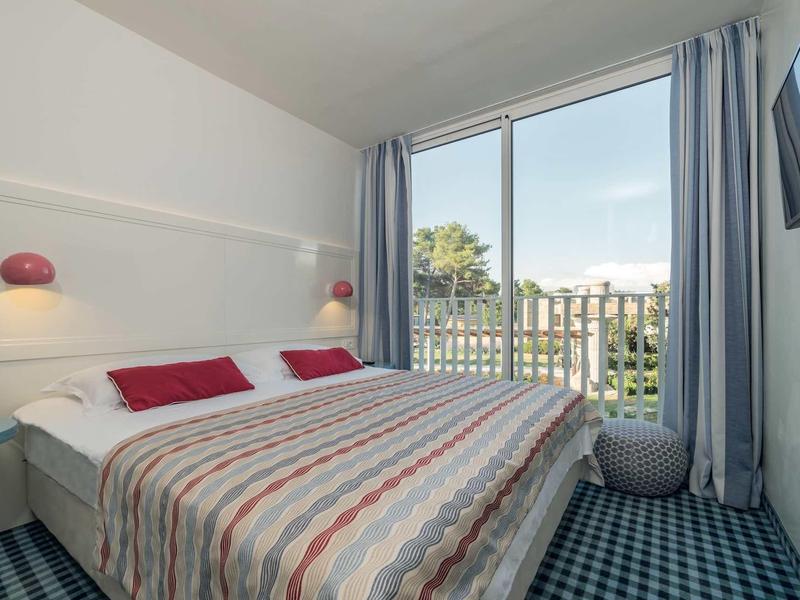 Lichte hotelkamer met tweepersoonsbed en balkon met uitzicht op bomen en stad.