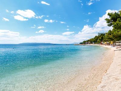 Klarer Strand mit türkisfarbenem Wasser und blauem Himmel, Bäume am rechten Rand.