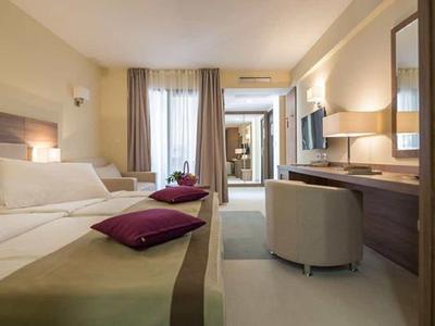 Hell eingerichtetes Hotelzimmer mit Doppelbett, Schreibtisch, Stuhl und Vorhängen am Fenster.