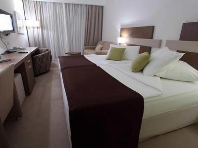 Modernes Hotelzimmer mit großem Doppelbett, braunen Tagesdecken und grünen Kissen, Schreibtisch und Fenster.