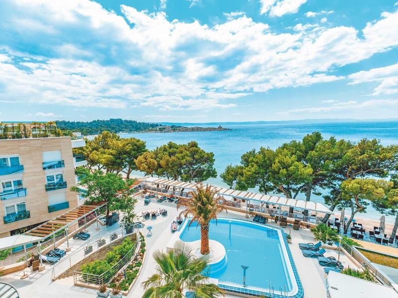 Moderner Gebäudekomplex mit blauem Pool, Palmen und grünen Bäumen am sonnigen Küstenufer.