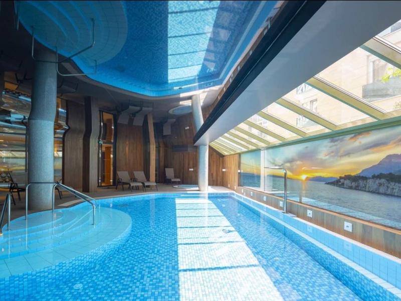 Innenpool mit blauem Wasser, Glasdach und großer Fensterfront mit Bergblick, moderne Architektur.