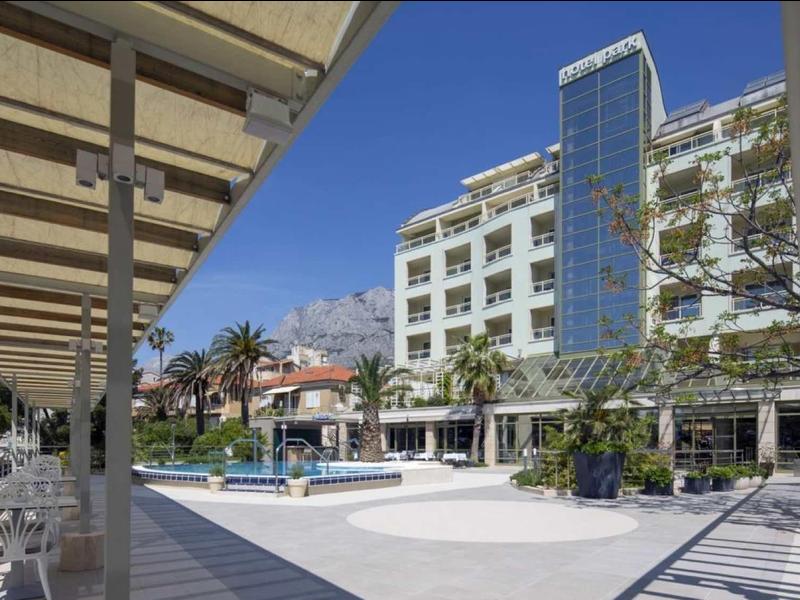 Moderner Hotelkomplex mit Pool, Sonnenterrasse, weißen Stühlen und blauem Himmel im Hintergrund.
