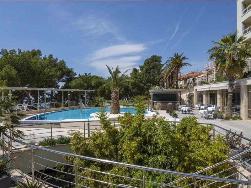 Moderner Außenpool mit Sonnenschirmen, Palmen und Liegestühlen neben einem Gebäude.