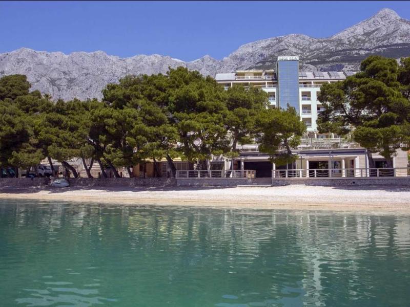 Klares türkisfarbenes Wasser vor einem Strand mit Bäumen und weißem Gebäude unter Bergen.