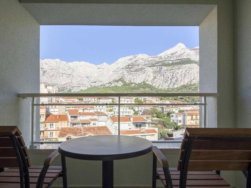 Balkon mit Tisch und zwei Stühlen, Blick auf Stadt und schneebedeckte Berge im Hintergrund.