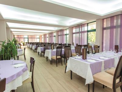 Jasna jadalnia z rzędami stołów i krzeseł, duże okna z fioletowymi zasłonami.