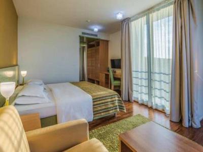 Nowoczesny pokój hotelowy z łóżkiem podwójnym, sofą, drewnianymi meblami i balkonem z zasłonami.