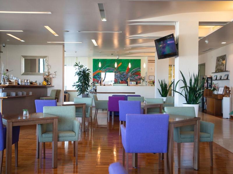 Nowoczesna restauracja hotelowa z kolorowymi krzesłami, drewnianą podłogą, roślinami i jasnym oświetleniem.