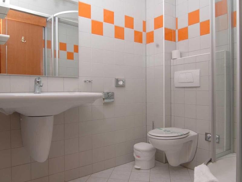 Kleines Bad mit weiß-orange Fliesen, Waschbecken, WC, Dusche und Spiegel mit Beleuchtung.